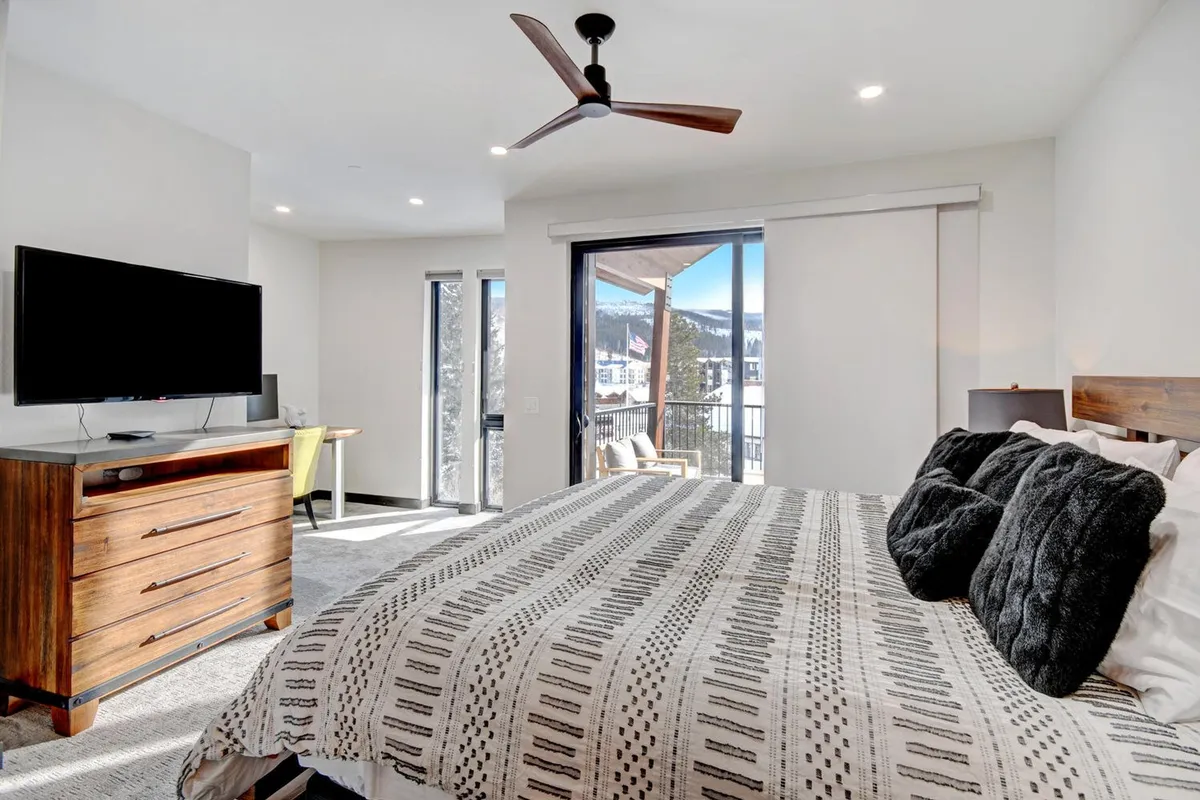 A serene bedroom featuring a plush bed with crisp bedding, a dresser, mounted TV, and access to a private balcony with scenic views. Natural light fills the space, creating an inviting and restful atmosphere.