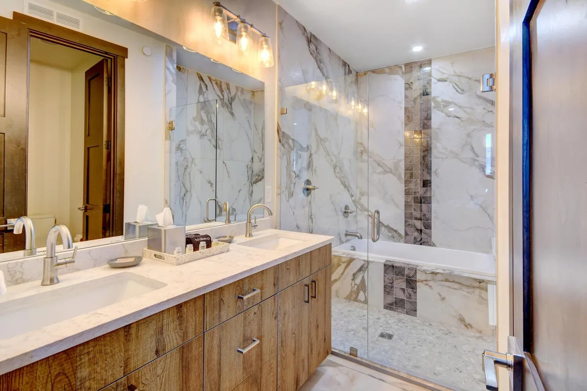 A luxurious ensuite bathroom featuring a double sink vanity, marble wall tiles, a glass shower enclosure, and a large mirror. The space is designed for relaxation and spa-like experiences.