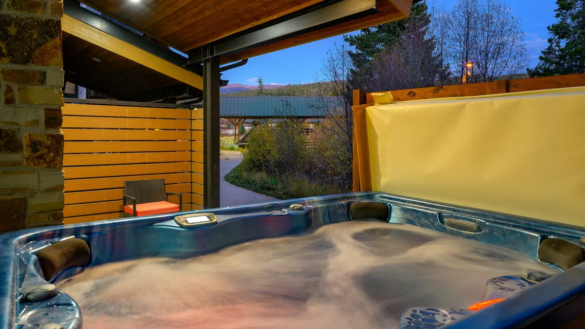 An exterior scene showcasing the hot tub surrounded by lush nature and a gentle pathway, with scenic mountain views in the background. The setting is tranquil and feels like a peaceful oasis, ideal for year-round use.