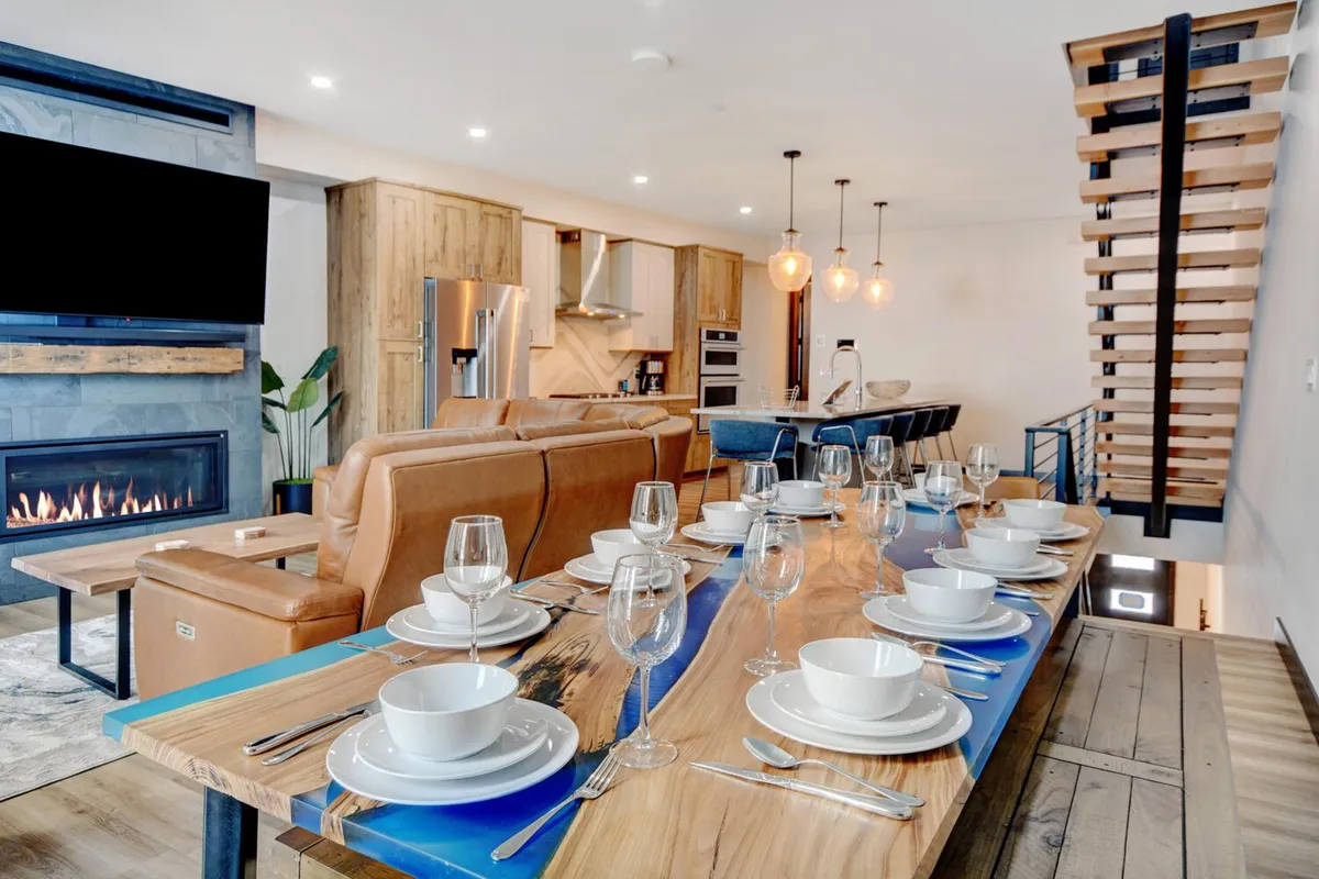An open-plan dining area set for a meal, featuring a dining table, modern staircase, fireplace, and a view into the kitchen. The atmosphere is warm and welcoming, with a sense of togetherness and celebration.