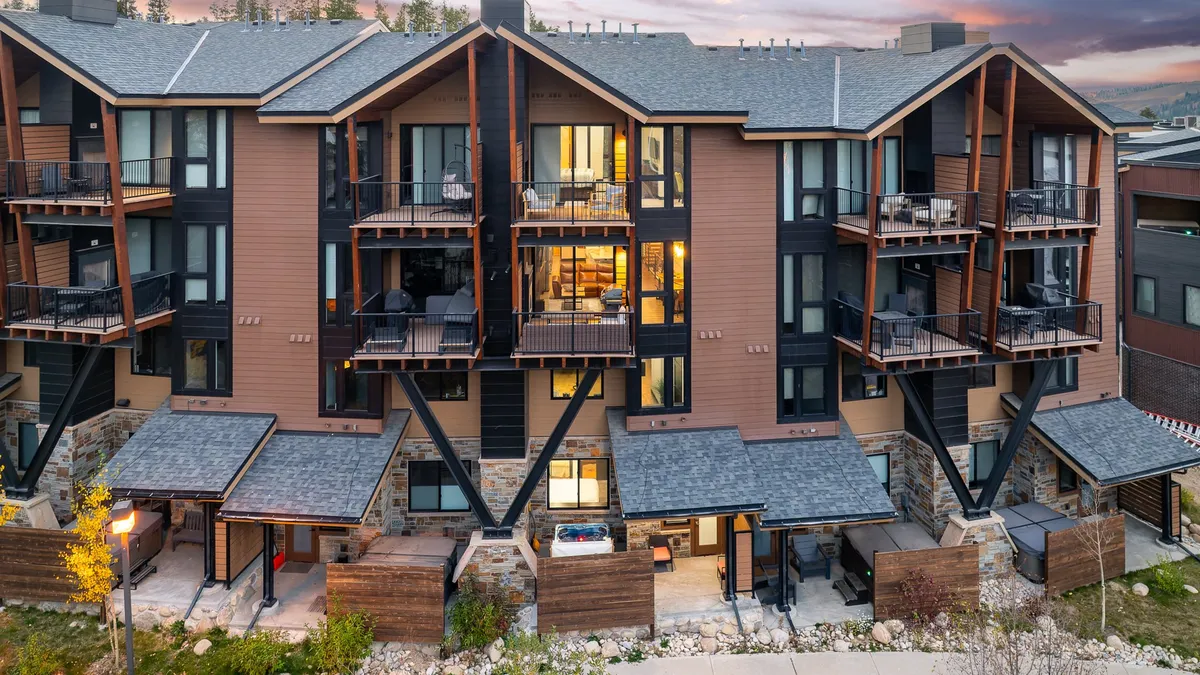 The image showcases the modern exterior of a luxury villa set against a Colorado sunset. Spacious balconies with inviting seating overlook a beautifully landscaped garden, while large windows hint at cozy interiors. Guests can be seen relaxing on the balconies and gathering around a fire pit, with the backdrop of Winter Park's scenic wilderness.
