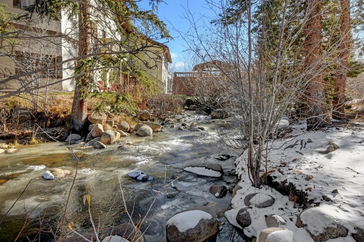 A serene outdoor scene with a flowing stream bordered by rock formations and greenery, with the building's facade visible beyond. The natural elements create a peaceful environment, inviting guests to connect with nature while enjoying the property's unique location next to Vasquez Creek.