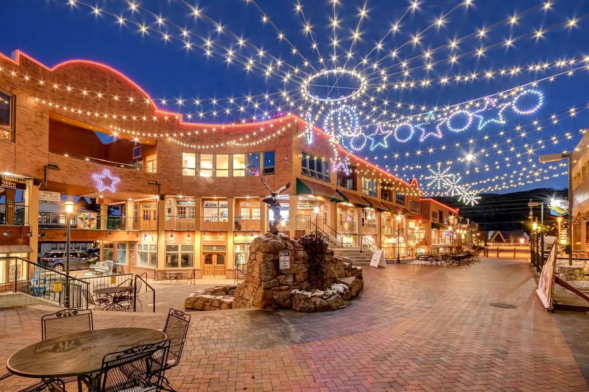 A lively outdoor scene featuring decorative string lights, a central fountain, brick pathways, and bustling storefronts. People are dining, shopping, or enjoying events, embodying the vibrant downtown experience regardless of season.