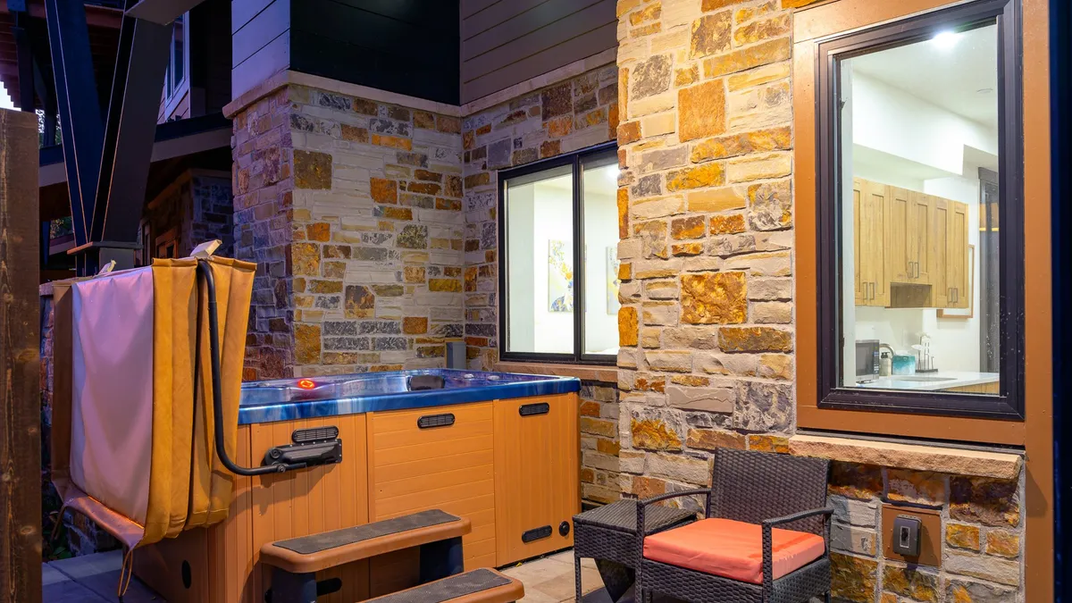 A tranquil exterior patio featuring a private hot tub nestled against a stone wall, with seating nearby and a window offering a peek into the cozy interior. Outdoor seating is perfectly positioned for socializing or quiet relaxation while enjoying views of surrounding nature.