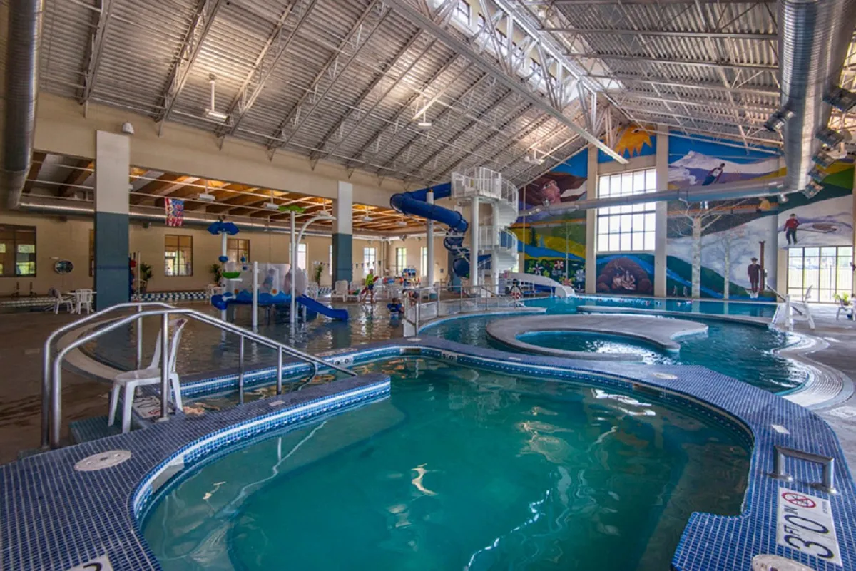 An energetic indoor recreation center with water slides, multiple pool areas, and colorful murals. Families and children can be seen swimming and playing, while others relax on poolside chairs. The lively, fun-filled atmosphere echoes the sense of resort-style amenities and recreation mentioned in the section.