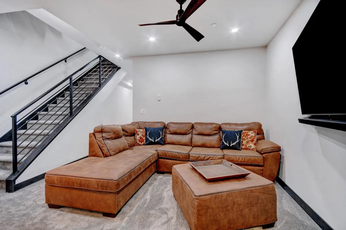 A spacious living area featuring a leather sectional sofa with decorative pillows, a modern coffee table, a large wall-mounted TV, and a staircase leading to another level. The room is styled in a comfortable, casual manner, ideal for relaxing, watching TV, and gathering with friends.