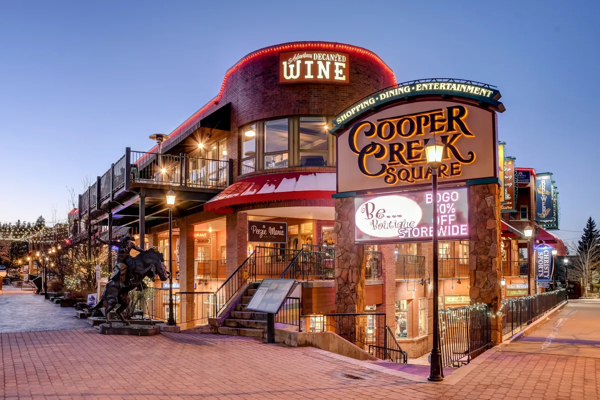 The Cooper Creek Square sign stands out amid a festive outdoor scene, with boutique storefronts, a wine shop, and decorative lighting, evoking the bustling, walkable downtown experience described in the section content.