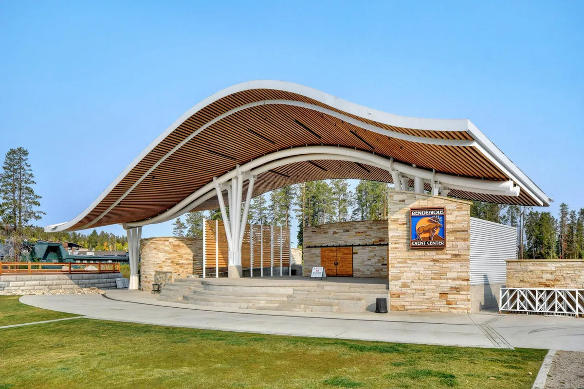 An open-air amphitheater stage with a curved wooden roof and a stone façade, set up for community events or concerts. The scene conveys a sense of local entertainment and gathering.