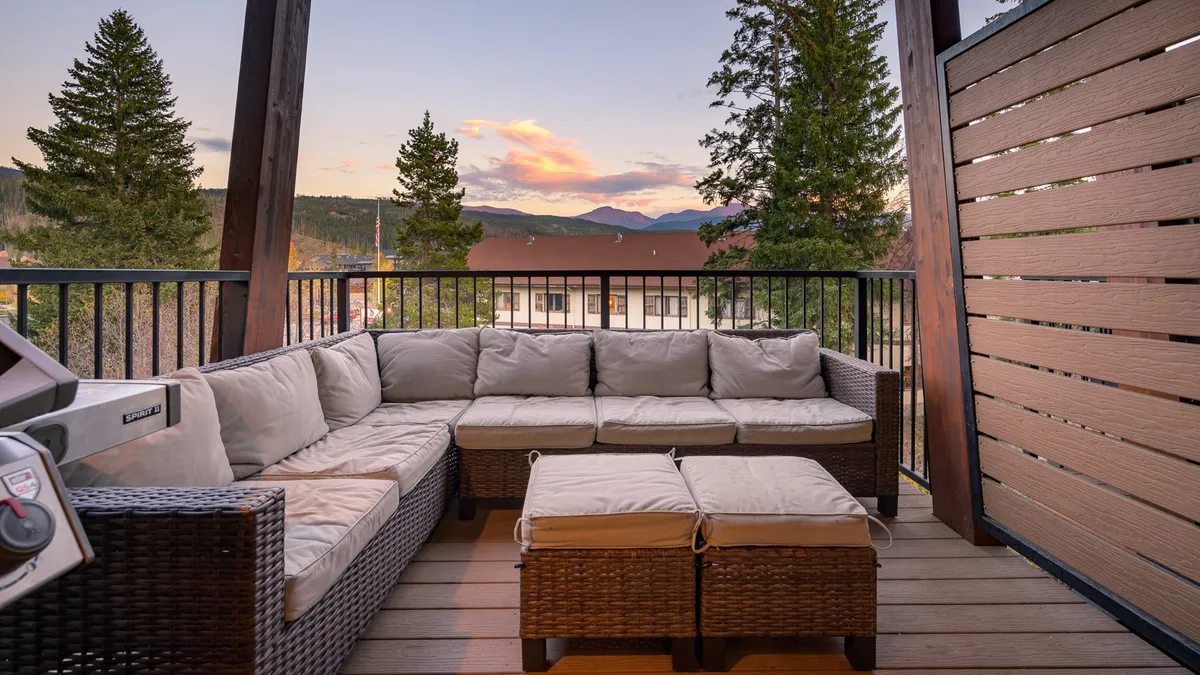 A spacious wooden deck features a cozy sectional sofa surrounded by lush trees and offers breathtaking mountain views. The outdoor space feels inviting, perfect for relaxing, entertaining, or gathering after a day's adventures. The setting sun casts a warm glow, adding to the sense of comfort and tranquility.