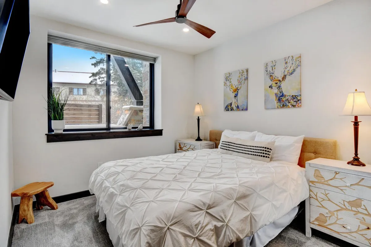 A tranquil queen bedroom with textured white bedding, colorful deer artwork above the headboard, matching nightstands with lamps, and natural light streaming through the window. The room feels peaceful and thoughtfully styled.